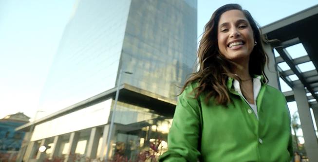 Atriz Camila Pitanga, de cabelos longos, sorri em frente a um prédio de vidro espelhado.