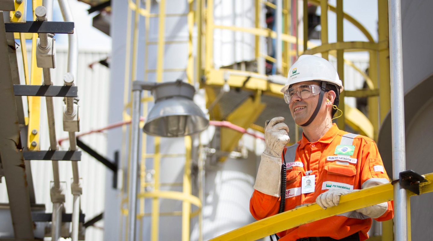 Petrobras professional wearing an orange uniform and PPE communicating by radio in an industrial area.