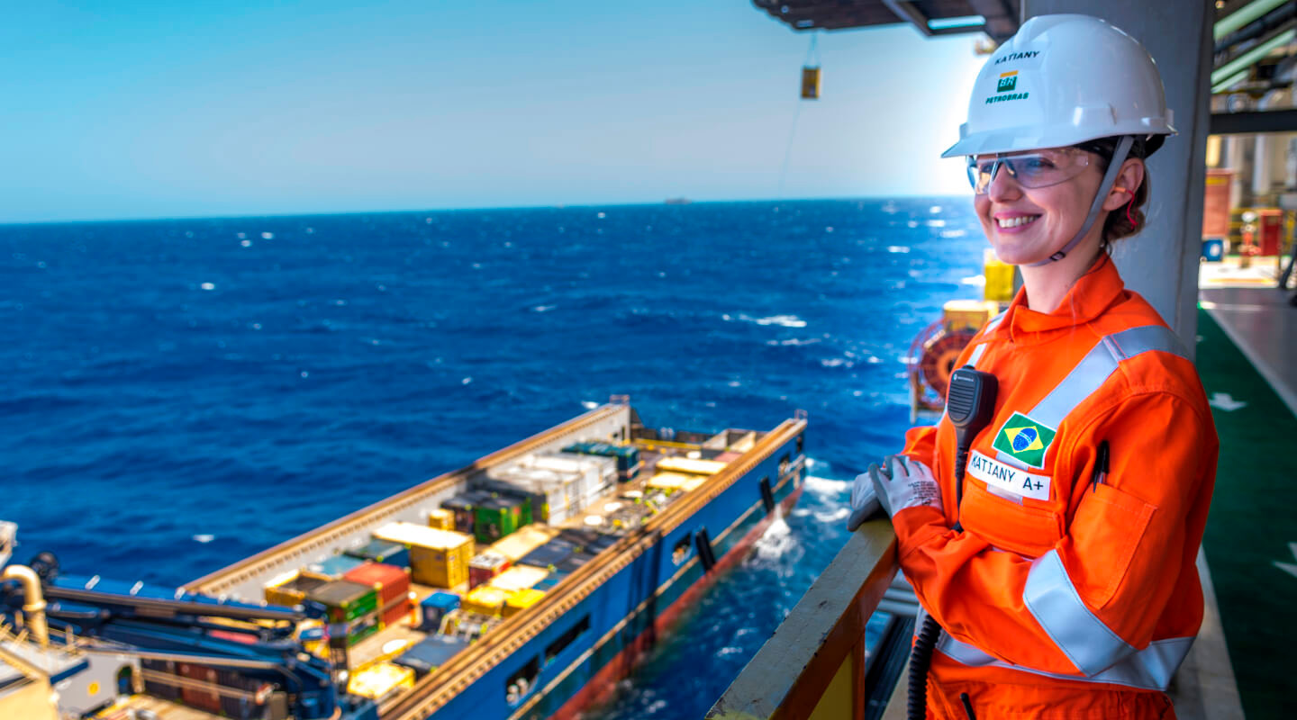 Petrobras worker in orange uniform and safety helmet smiling on offshore platform with the ocean and supply vessel in the background.